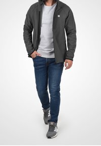 Graue Softshell-Jacke mit Reißverschluss vorne, Taschendetails, kombiniert mit einem grauen Sweatshirt und blauen Jeans. Die Sneakers haben graue und weiße Akzente.