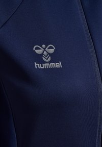 Marinblå sportjacka av slät tyg med dragkedja och en silverlogga på vänster bröst.