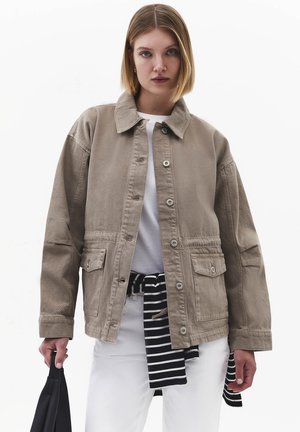 Junge Frau mit kurzen blonden Haaren, die eine beige Jacke mit Knöpfen über einem weißen Hemd, weißen Hosen trägt und eine schwarze Tasche in einer Hand hält.