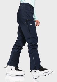 Marineblaue Skihosen mit einer figurschmeichelnden Passform, ausgestattet mit Reißverschlusstaschen und verstärkten unteren Säumen. Kombiniert mit weißen Skischuhen und einem türkisfarbenen Oberteil.