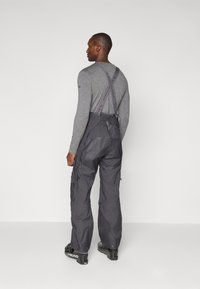 Graues Langarmshirt unter dunkelgrauen Latzhosen mit verstellbaren Trägern. Bemerkenswerte Hardware-Details an den Hosenträgern und seitlichen Taschen. Schwarze Stiefel.