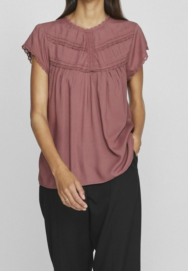 Blusa a maniche corte di un delicato rosa con rifiniture in pizzo e un design di tessuto arricciato. Indossata con pantaloni neri, che evidenziano una vestibilità loose e una consistenza morbida.
