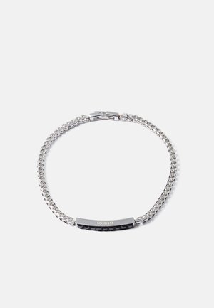 Bracelet en chaîne argentée avec une barre rectangulaire gravée "GUESS" et des clous pyramides noirs sur un fond blanc.