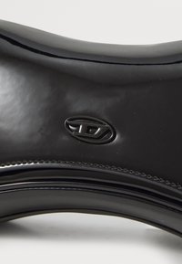 Scarpa nera lucida con un design elegante e una forma curva, caratterizzata da un logo ben visibile e una superficie liscia.
