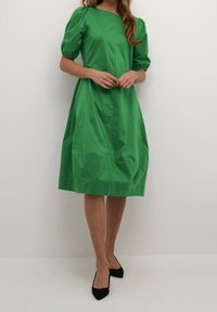 Grön bomullsklänning med korta puffärmar, rund halsringning, ledig passform och knälång fåll, parat med svarta spetsiga skor.