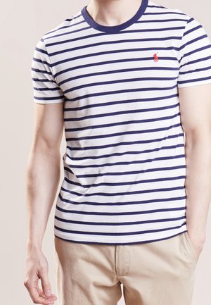 Uomo che indossa una maglietta bianca con righe orizzontali blu navy e un logo rosso sul petto, pantaloni beige, in piedi con una mano in tasca.