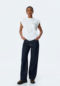 Hvid bomulds-sleeveless t-shirt med lomme og sølv kædedetalje, parret med mørkeblå vide denim jeans. Brune sko.