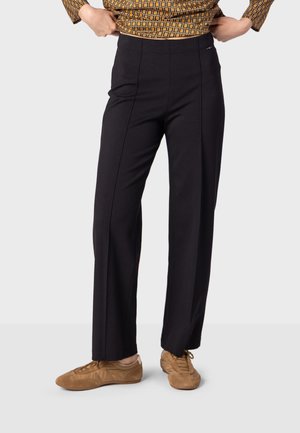 Personne portant un pantalon noir à jambes droites associé à des baskets marron à lacets et un haut à manches longues à motif, debout, les mains ajustant la taille.