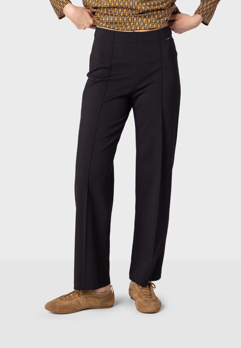 Personne portant un pantalon noir à jambes droites associé à des baskets marron à lacets et un haut à manches longues à motif, debout, les mains ajustant la taille.