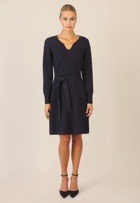 Abito lavorato a maglia blu navy con scollo a V, maniche lunghe e cintura annodata in vita. Tessuto liscio, lunghezza al ginocchio, abbinato a tacchi neri.