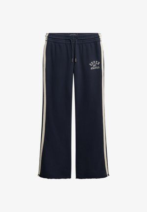 Pantaloni da tuta larghi blu navy con strisce bianche ai lati, fascia elasticizzata in vita, cordoncino e logo "Super Dry Athletics" sulla coscia sinistra.