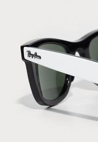Mustat ja valkoiset aurinkolasit paksulla kehyställä, vihreillä linsseillä ja hopeisella Ray-Ban-logolla templeissä. Sileä pinta, retro muotoilu.
