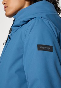 Profil osoby v modrej mikine s kapucňou a čiernym zipsom, na rukáve s čiernou nášivkou s nápisom "ICEPEAK".