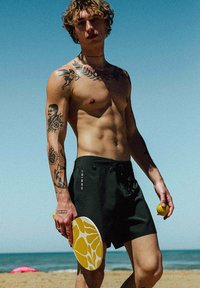 Shorts de bain noirs avec un cordon de serrage, tenant une balle jaune et une pagaie avec un motif abstrait blanc et jaune, en arrière-plan une plage.