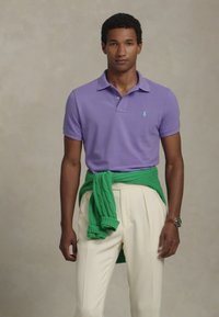 Moški v svetlo vijoličnem polo majici, kremastih hlačah in z zelenim puloverjem, zavezanem okoli pasu, stoji pred enobarvno podlago.