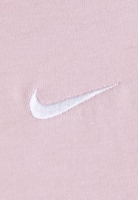 Ljusrosa tyg med en mjuk textur, prydligt med en broderad vit Nike swoosh-logga. Designen är minimalistisk och fokuserar på loggans detalj.