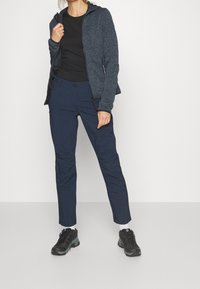 Mörkblå åtsittande byxor kombinerade med en texturerad kolgrå jacka med huva, blixtlåsfickor och en svart t-shirt under.