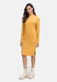 Frau mit langen, dunklen Haaren, die ein senfgelbes geripptes Strickkleid und cremefarbene Stiefeletten trägt und vor einem schlichten Hintergrund steht.