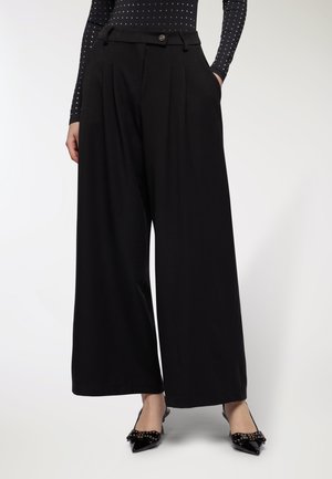 Donna che indossa pantaloni neri a vita alta e a gamba larga, abbinati a ballerine nere a punta con dettagli di fiocchi borchiati.