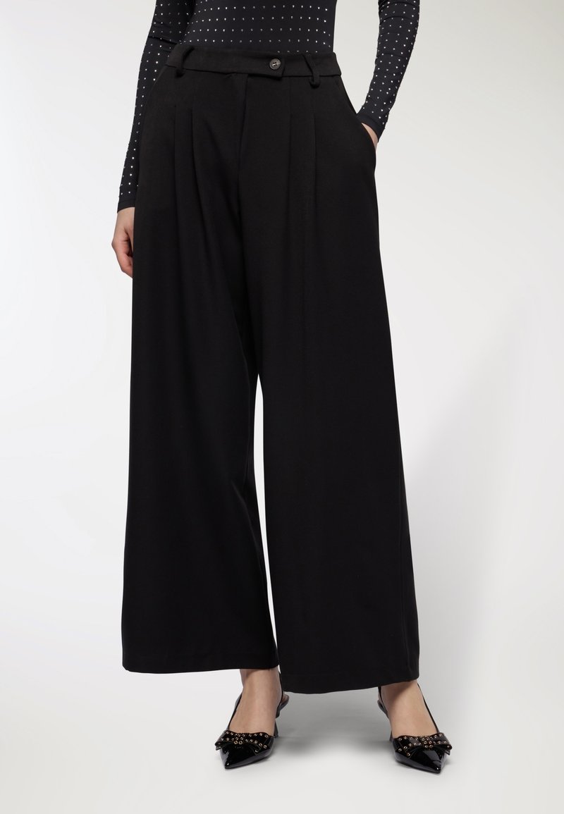 Donna che indossa pantaloni neri a vita alta e a gamba larga, abbinati a ballerine nere a punta con dettagli di fiocchi borchiati.