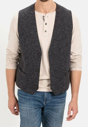 Mężczyzna w ciemnoszarej kamizelce zapinanej na guziki, na beżowej koszulce typu henley z długim rękawem i niebieskich dżinsach, stojący z rozluźnionymi ramionami.