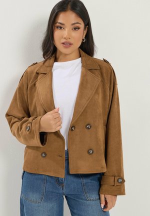 Woman wearing a brown double-breasted jacket over a white shirt and blue jeans, standing against a plain light background.