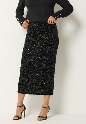 Zwarte midi-rok met pailletten gedragen met zwart langermige top en zwarte hoge hakken sandaal met enkelriem op een lichte achtergrond.