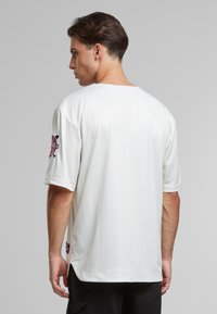 Maillot blanc à manches courtes avec des rayures verticales, encolure ronde, fentes sur les côtés et un écusson logo coloré sur la manche gauche. Texture lisse.