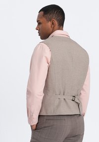 Chaleco de tela gris con textura suave, correa trasera ajustable, usado sobre una camisa de manga larga rosa claro, combinado con pantalones de cuadros.
