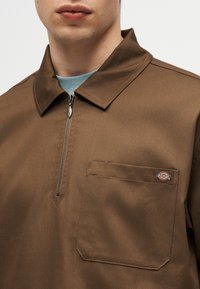 Άνδρας που φοράει καφέ πουκάμισο με φερμουάρ, γιακά και τσέπη στο στήθος με μικρό λογότυπο Dickies.