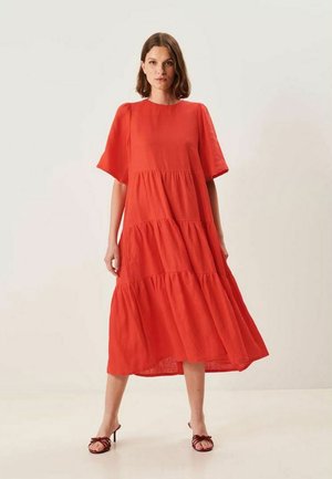 Kvinde iført løs, lagdelt, knaldrød midikjole med korte ærmer og brune sandaletter med hæl, stående på en ensfarvet baggrund.