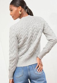 Szary sweter dzianinowy z warkoczowym wzorem, ściągaczami na mankietach i u dołu. W połączeniu z niebieskimi dżinsami, prezentującymi dopasowany krój i zarys sylwetki.