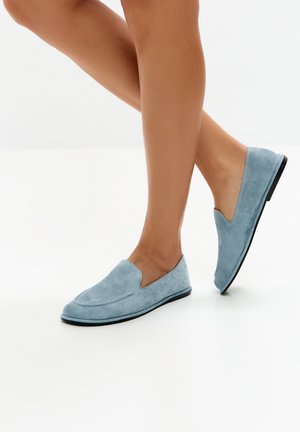 Persona che indossa mocassini slip-on in camoscio azzurro chiaro con suola nera, in piedi su una superficie bianca con un piede leggermente sollevato.