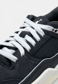 Baskets en daim noir avec des lacets blancs, un panneau en mesh et un accent de logo près de la languette. La semelle présente une couleur claire contrastante le long du bord.