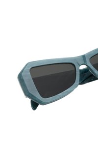 Un paio di occhiali da sole angolari realizzati in plastica traslucida color teal con lenti grigie scure e un design unico e sfaccettato.