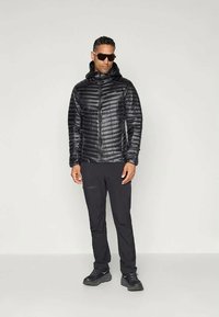 Άνδρας φορώντας μαύρο μπουφάν με κουκούλα, μαύρο παντελόνι, μαύρα αθλητικά παπούτσια και σκούρα γυαλιά ηλίου, να στέκεται σε απλή ανοιχτόχρωμη φόντο.