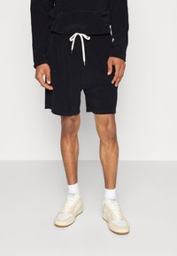 Shorts en tissu éponge noir avec taille à cordon, associés à des chaussettes blanches et des baskets beiges. Le modèle est debout devant un fond neutre.