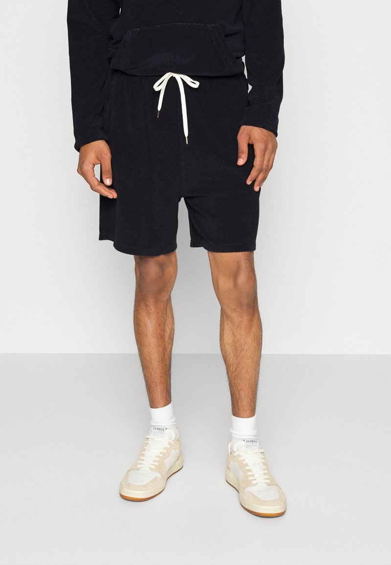 Shorts en tissu éponge noir avec taille à cordon, associés à des chaussettes blanches et des baskets beiges. Le modèle est debout devant un fond neutre.