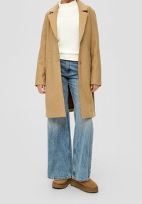 Długi płaszcz camelowy na kremowym swetrze z dzianiny, w zestawie z szerokimi niebieskimi dżinsami i brązowymi botkami za kostkę. Minimalistyczny design z wyraźnymi liniami.