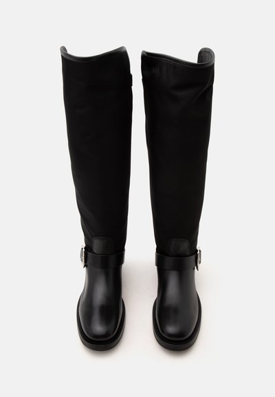 Bottes hautes en cuir noir avec des bouts arrondis et des détails avec des sangles près de la cheville, debout sur un fond blanc.