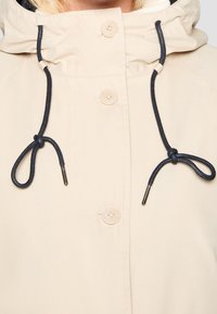 Veste à capuche beige avec une doublure douce, dotée de deux grands boutons et d'un cordon noir réglable au niveau du cou.