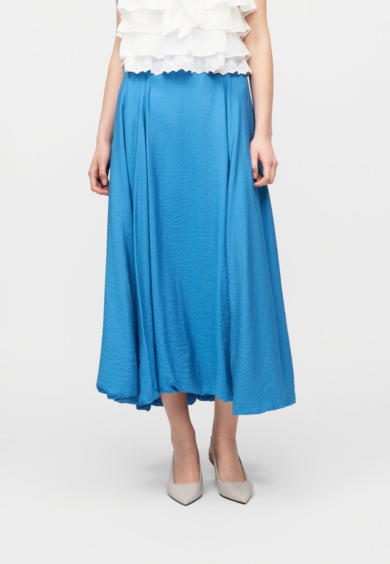 Γυναίκα που φοράει φωτεινή μπλε midi φούστα με σούρα στο κάτω μέρος, σε συνδυασμό με λευκό πουκάμισο με βολάν και γκρι παπούτσια με μυτερή μύτη.