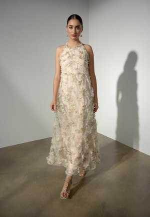 Frau trägt ein ärmelloses, knöchellanges, transparentes Blumenmusterkleid mit Rüschendetails und hochhackigen Riemchensandalen, steht in Innenräumen.