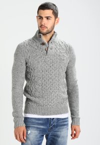 Suéter gris de punto con un patrón de trenzado, cuello alto y botones de madera. Combinado con jeans azul desgastados, mostrando un estilo casual.