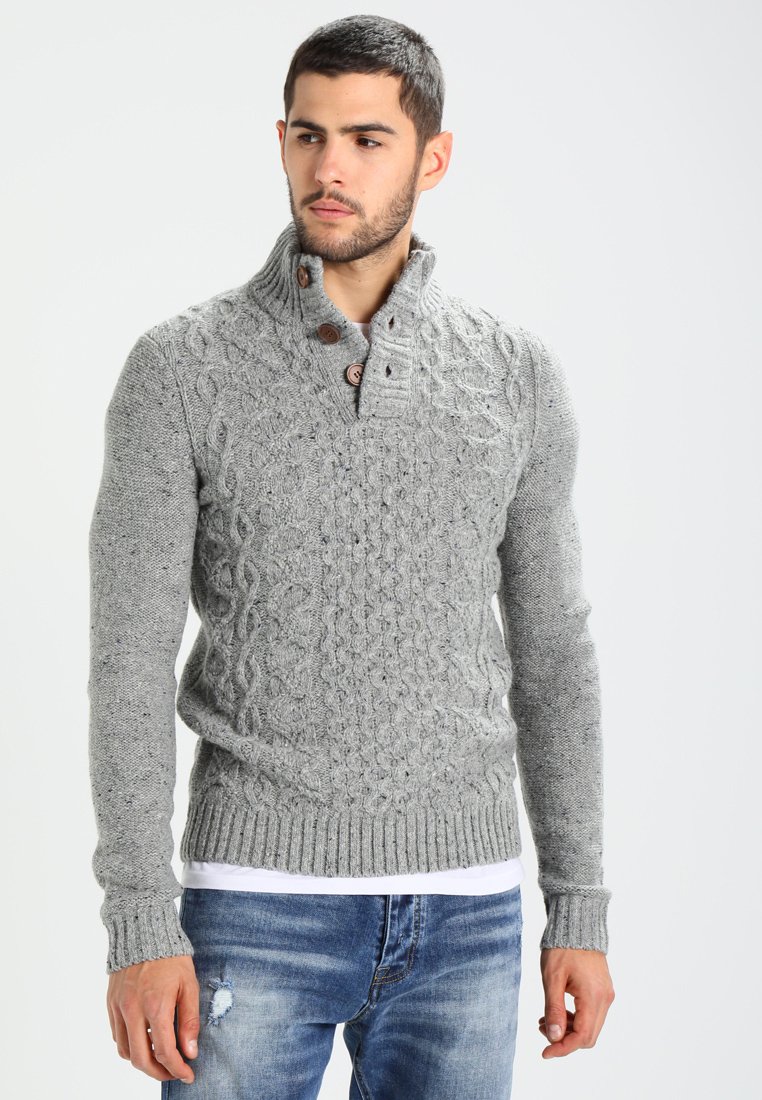 Suéter gris de punto con un patrón de trenzado, cuello alto y botones de madera. Combinado con jeans azul desgastados, mostrando un estilo casual.