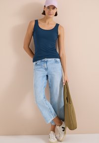 Mujer con una camiseta sin mangas azul marino, jeans azul claro recortados, zapatillas blancas y una gorra rosa, sostiene una chaqueta verde oliva con una mano.