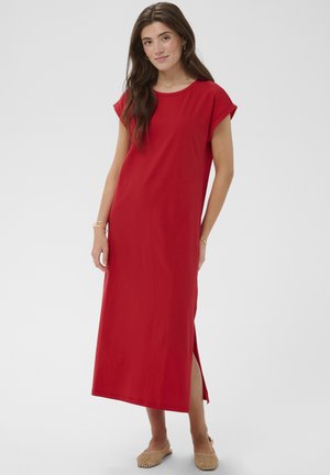 Young woman with long dark hair wearing a loose red ankle-length dress with short sleeves and beige woven flats.