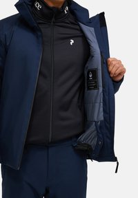Námorně modrá bunda se strukturovaným designem, s hladkým zipem a vnitřním podšívem. Černá přiléhavá základní vrstva pod ní, zobrazující detaily značky.