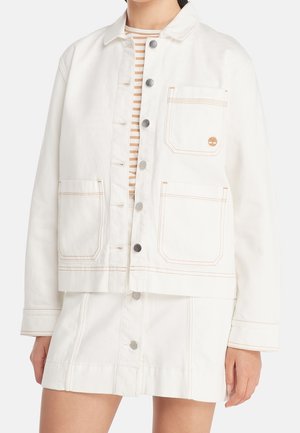 Kvinde iført en off-white denimjakke med knapper og tre forlommer samt matchende nederdel, kombineret med en beige og hvid stribet top.