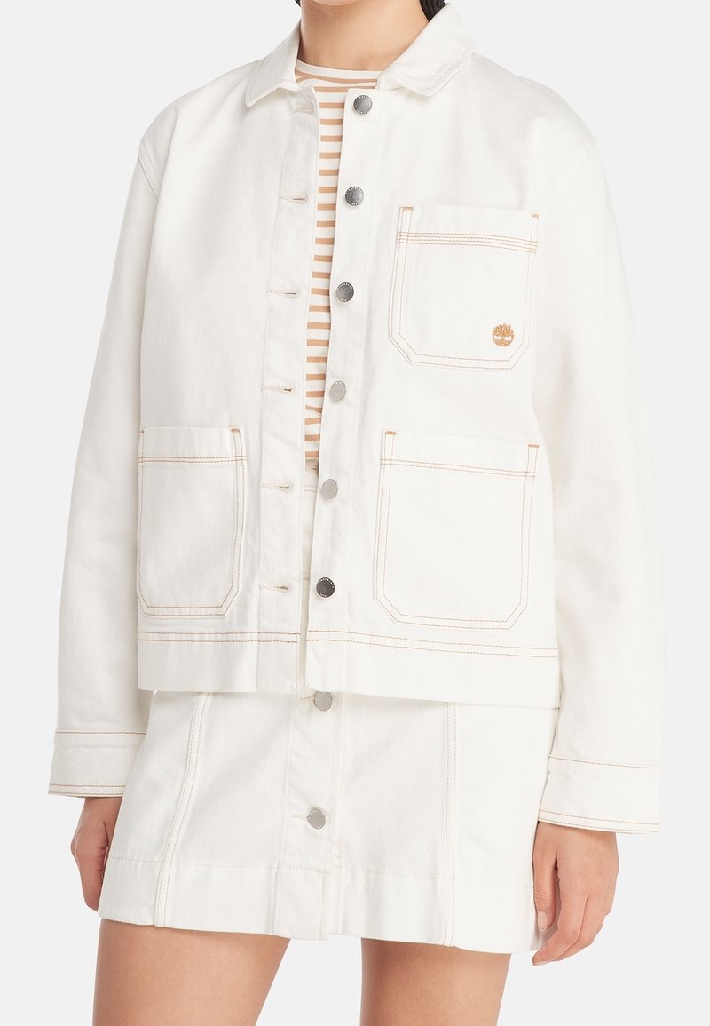 Femme portant une veste en jean écrue boutonnée avec trois poches à l'avant et une jupe assortie, associée à un top rayé beige et blanc.